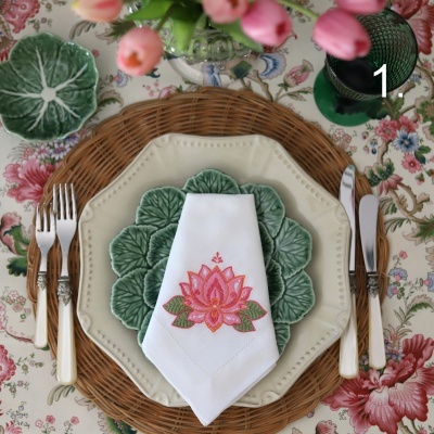 Mesa posta com pratos cerâmicos creme e verdes, guardanapo branco bordado e talheres variados sobre toalha floral.