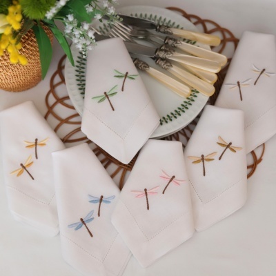 Guardanapos bordados com libélulas coloridas ao redor de prato com talheres em fundo branco.