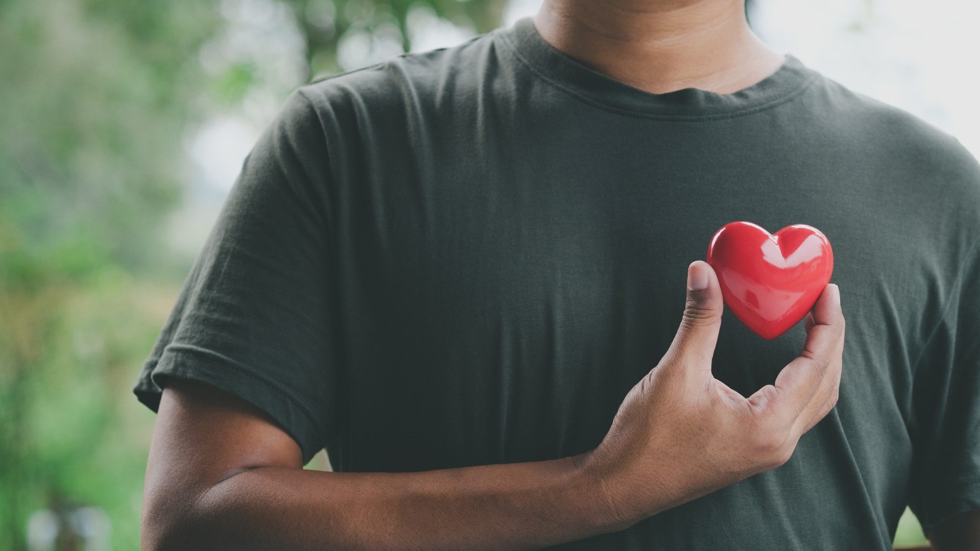Pessoa com t-shirt cinzenta escura segurando coração vermelho brilhante perto do peito