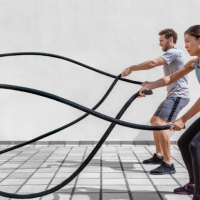 Homem e mulher a fazer exercício com cordas de batalha num espaço com piso cerâmico e parede branca.