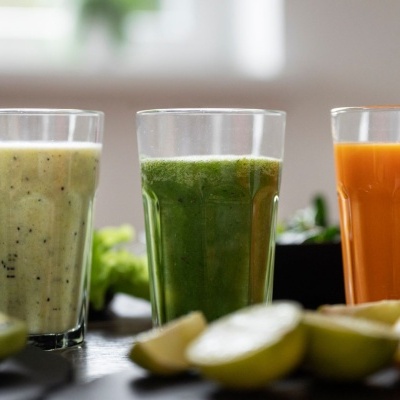 Três copos com sumos de cores diferentes em mesa com frutas e vegetais ao fundo