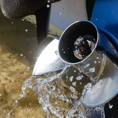 Hélice metálica prateada com água a cair, fixada a motor azul numa água rasa