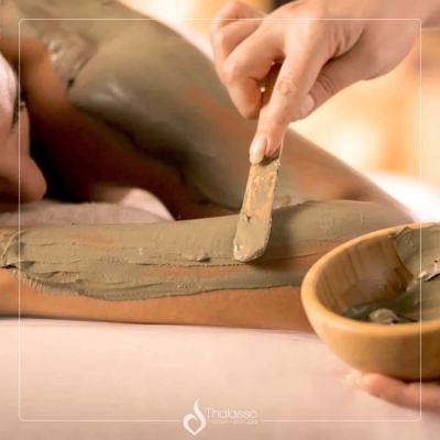 Aplicação de máscara de lama verde no braço com espátula e tigela de madeira ao lado