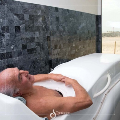 Homem idoso relaxa numa cápsula branca de hidroterapia com parede cinzenta azulada e janela de vidro.