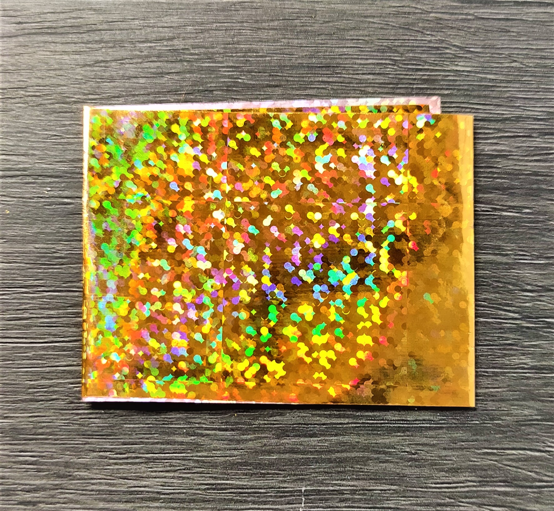 Envelope holográfico dourado com reflexos coloridos sobre superfície cinzenta texturada
