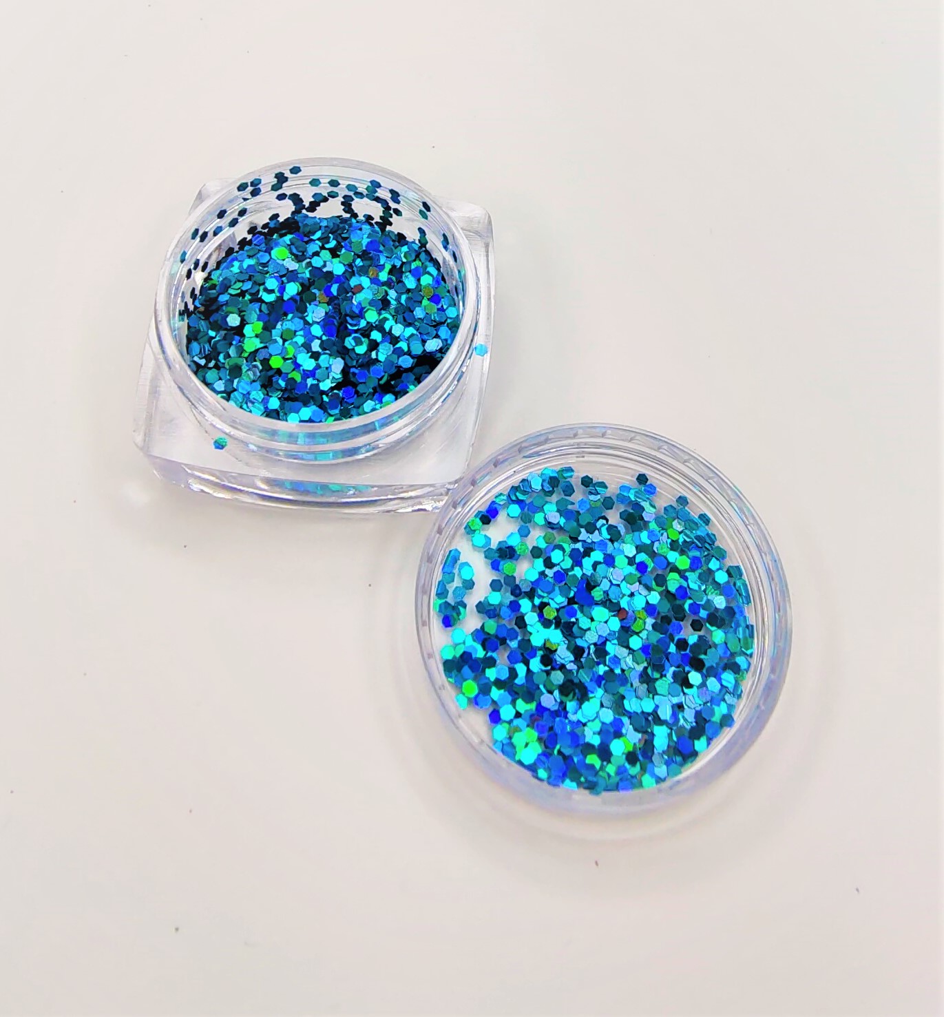 Dois frascos pequenos transparentes com glitter azul verde prateado sobre superfície branca