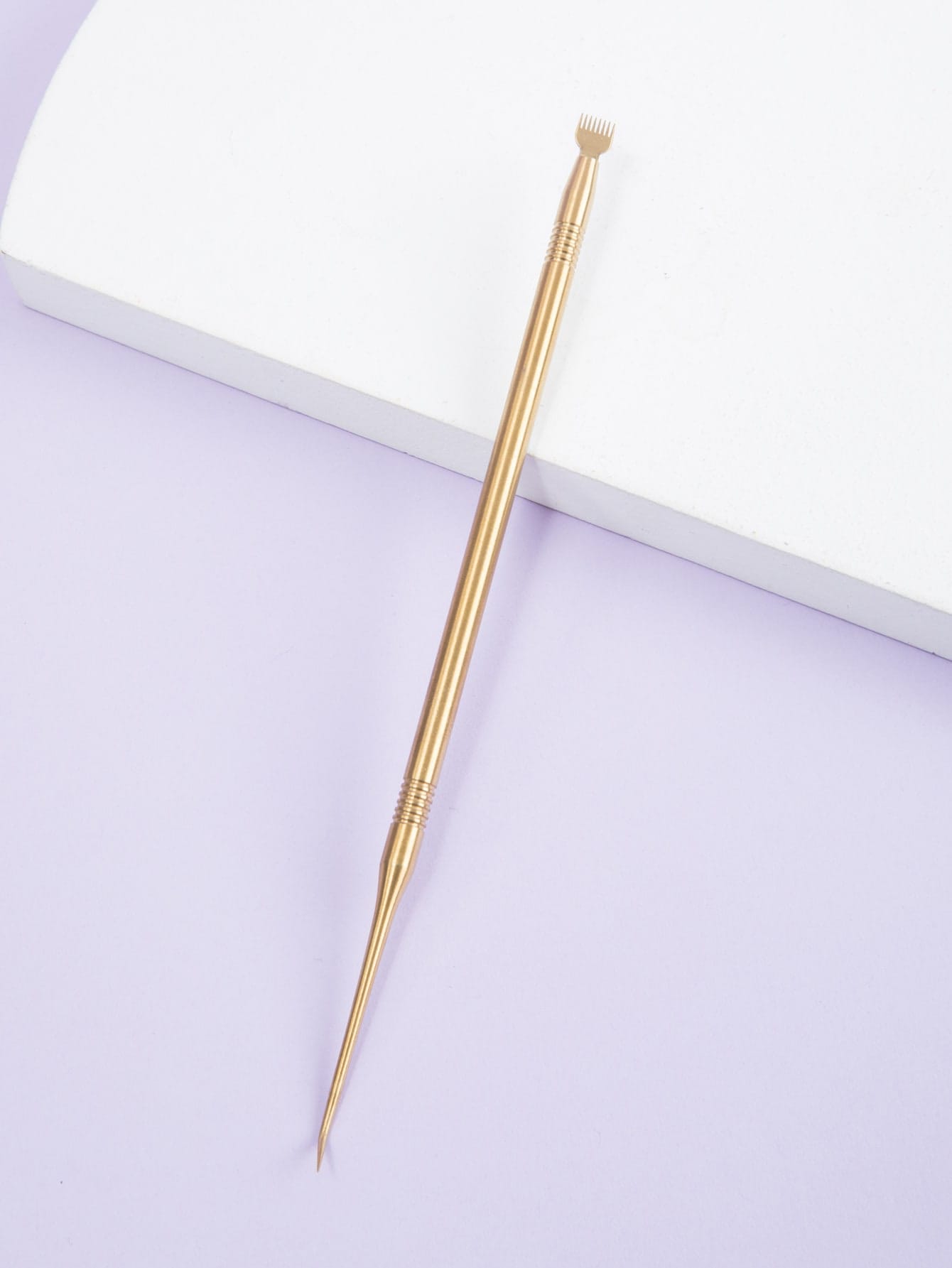 Ferramenta metálica dourada com ponta de pente e ponta fina sobre fundo branco e lilás