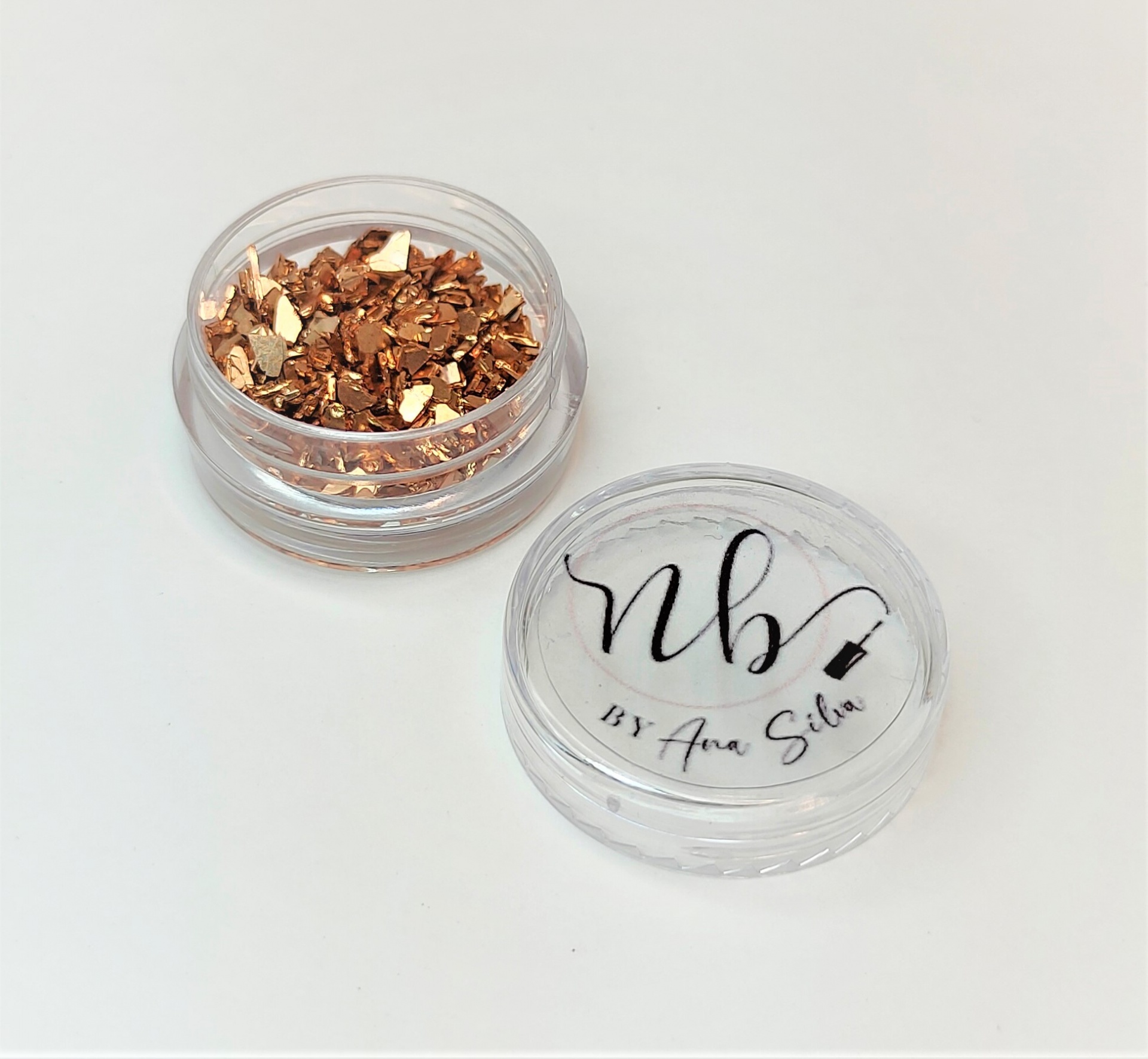 Pote transparente com pedaços metálicos dourados e tampa com texto 'nb BY Ana Silva'.