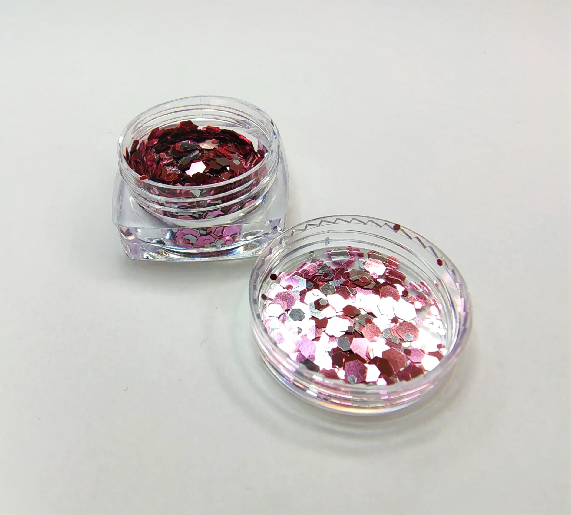 Duas caixas pequenas transparentes com glitter hexagonal rosa, vermelho e branco sobre fundo branco.