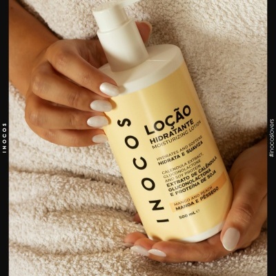 Frasco amarelo de loção hidratante INOCOS com bomba, segurado por mãos com unhas brancas, sobre tecido branco.