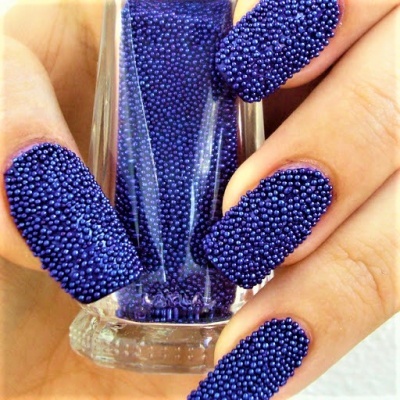 Unhas decoradas com esferas azuis em textura tridimensional segurando um frasco transparente com esferas azuis