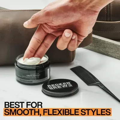 Mão pega pomada de cabelo num frasco preto com pente e bolsa marrom ao fundo