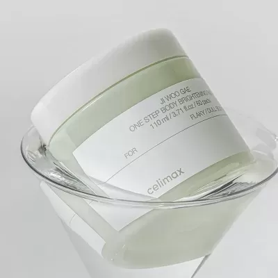 Frasco de creme corporal verde com tampa branca e rótulo branco inclinado numa taça de vidro.