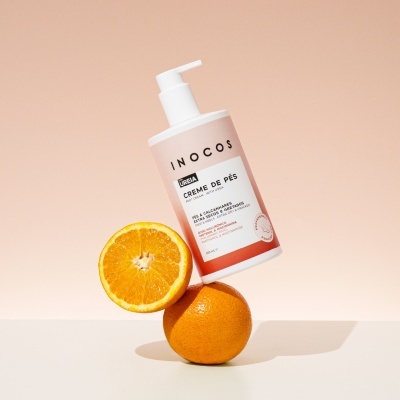 Frasco de creme de pés INOCOS com doseador branco e tangerinas sobre fundo bege claro.