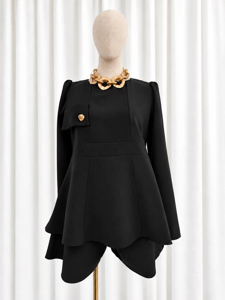 Conjunto Nancy em preto Fato feminino preto com saia assimétrica, manga comprida, bolso com botão dourado e colar dourado em manequim frente a cortinas brancas
