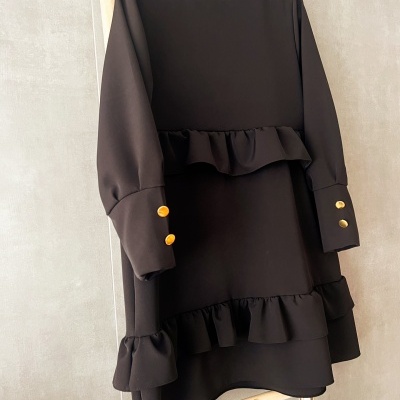 Vestido preto de mangas compridas com folhos e botões dourados pendurado num cabide.