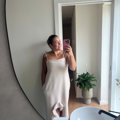 Vestido comprido bege claro com alças finas e detalhe franzido na bainha refletido num espelho redondo.