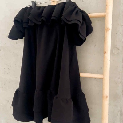 Vestido preto com folhos pendurado numa escada de madeira contra parede cinzenta.