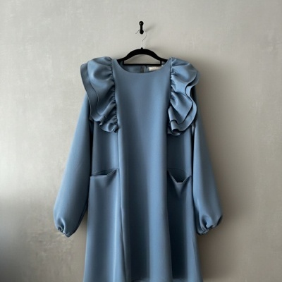 Vestido azul claro com folhos nos ombros e bolsos, pendurado numa parede cinzenta