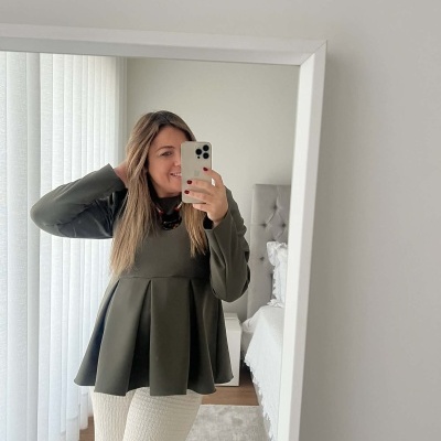 Mulher com blusa verde escuro e calças brancas a tirar selfie em espelho num quarto.