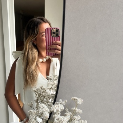 Mulher com vestido branco e colar de pérolas a tirar selfie no espelho oval com flores brancas à frente.