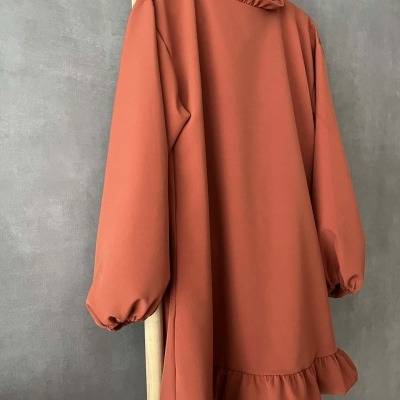 Vestido terracota de manga comprida com folhos na gola, punhos e barra pendurado num cabide preto numa estrutura de madeira.