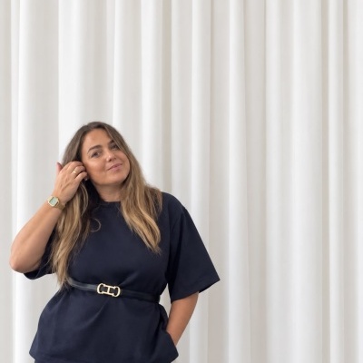 Mulher com vestido preto e cinto posando em frente a cortina branca