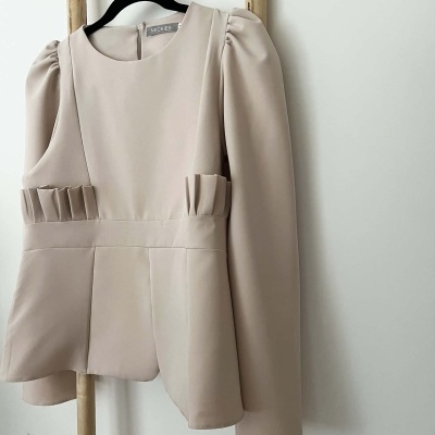 Blusa bege claro com mangas compridas e franzidos na cintura pendurada em cabide preto numa estrutura de madeira