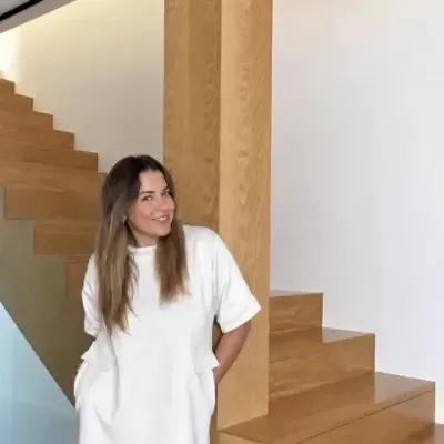 Mulher com vestido branco curto e mangas curtas junto a escadas e pilares de madeira clara