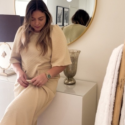 Mulher com vestido bege sentada junto a mesa com vaso e espelho circular na parede