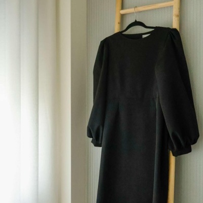 Vestido preto de manga comprida pendurado numa estrutura de madeira junto a uma janela com cortinas brancas.