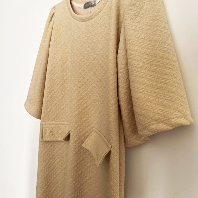 Vestido bege claro com mangas compridas e bolsos, textura quadriculada, pendurado em cabide preto.