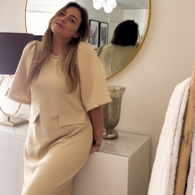 Vestido bege texturizado feminino com bolsos falsos, mulher junto a móvel branco e espelho redondo