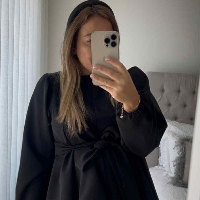 Pessoa com blusa preta de manga comprida com laço na cintura tirando uma selfie no quarto