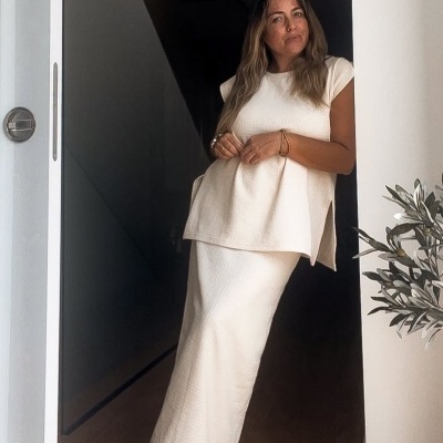 Mulher vestindo vestido longo creme e blusa sem mangas creme em interior com chão de madeira.