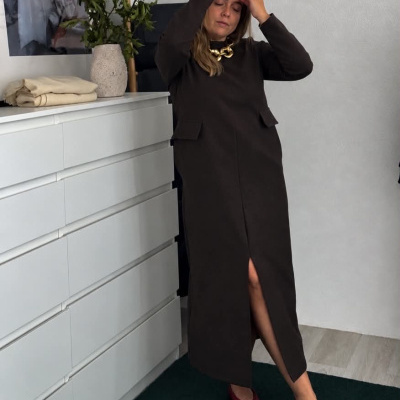 Mulher com vestido comprido castanho escuro e sapatos vermelhos junto a cómoda branca com vaso e roupas dobradas