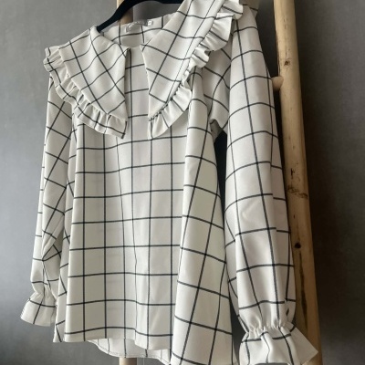 Blusa xadrez branca com gola e folhos pendurada em cabide sobre escada de madeira