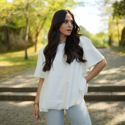 Mulher com blusa branca de manga curta e calças cinza num parque com árvores.