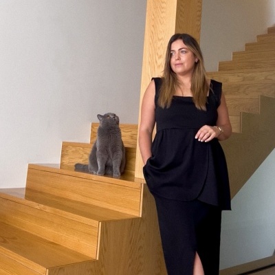 Mulher com vestido preto ao lado de escada de madeira clara com gato cinzento sentado nas escadas