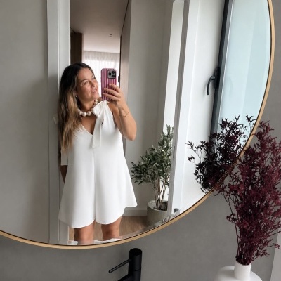 Mulher com vestido branco a tirar selfie em espelho redondo num banheiro moderno.