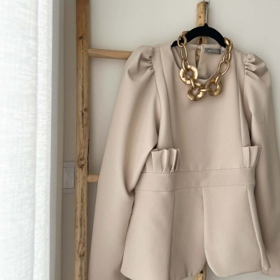 Blazer bege com mangas franzidas e colar dourado pendurado em estrutura de madeira.