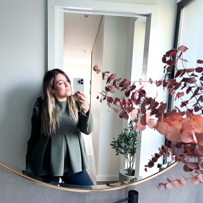 Espelho redondo refletindo mulher com blusa verde e ramos secos cor de tijolo em vaso