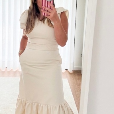 Vestido comprido creme com mangas bufantes e saia com pregas num interior com chão de madeira e cortinas brancas