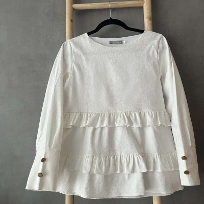 Blusa branca texturizada com folhos e botões, pendurada numa escada de madeira