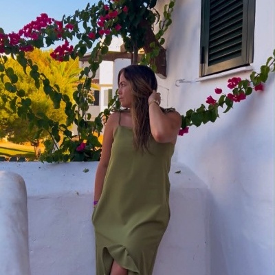 Vestido longo verde musgo com folhos usado por mulher ao ar livre junto a parede branca e flores vermelhas