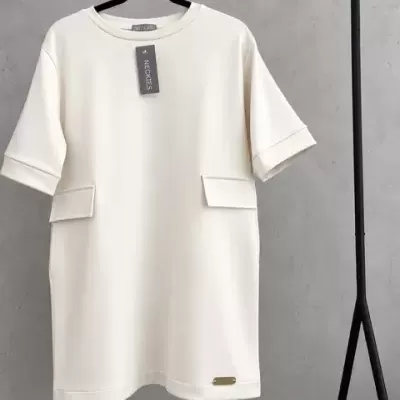 Vestido branco creme com bolsos, etiqueta NECKTIES e placa metálica pendurado em suporte preto.