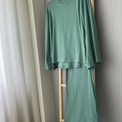 Conjunto de roupa verde claro com camisola e calças pendurado em suporte de madeira junto a cortinas brancas