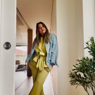 Mulher vestida com camisa de ganga azul e conjunto amarelo com nó frontal ao lado de planta em interior.