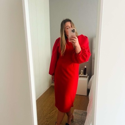 Mulher veste vestido vermelho com mangas volumosas a tirar selfie num espelho num quarto com cama e criado-mudo.
