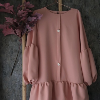 Blusa rosa claro com botões e folhos pendurada numa escada de madeira com ramo de folhas roxas.